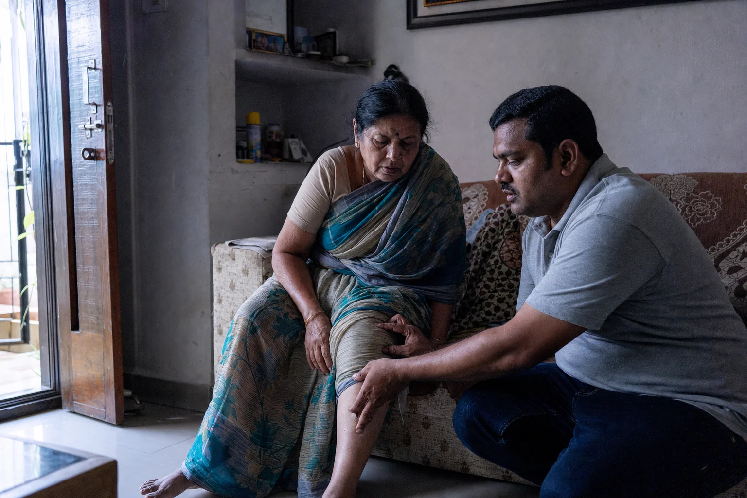 A physiotherapist crouched beside an older woman seated on a sofa in a Hyderabad flat, both looking at her knee.