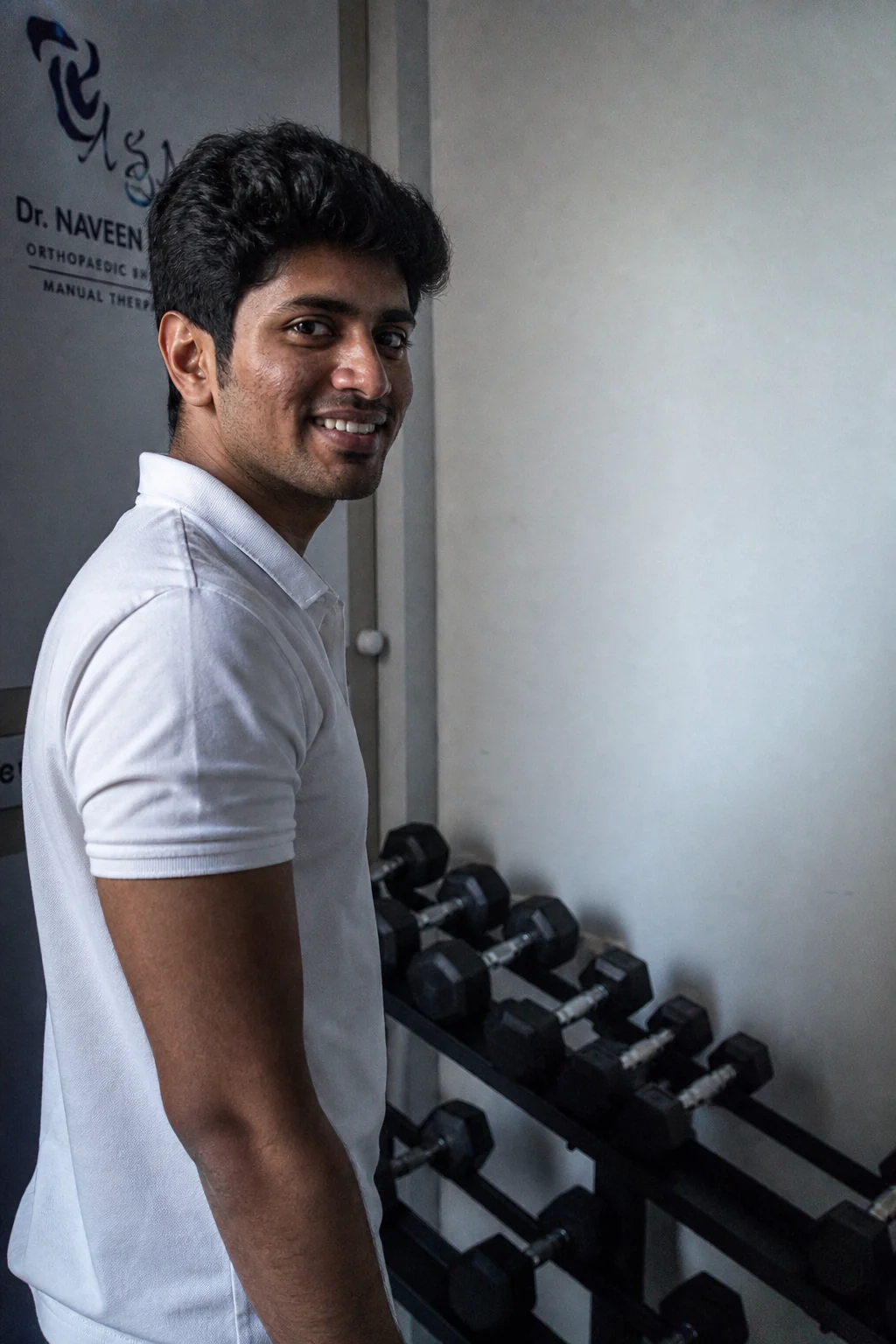 Dr Naveen Gudipati beside a rack of dumbbells, turned back over his shoulder towards the camera.